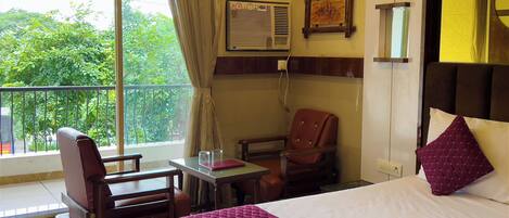 A hotel room with a bed, a desk, a chair, a window with a view of greenery, and a wall-mounted air conditioning unit.
