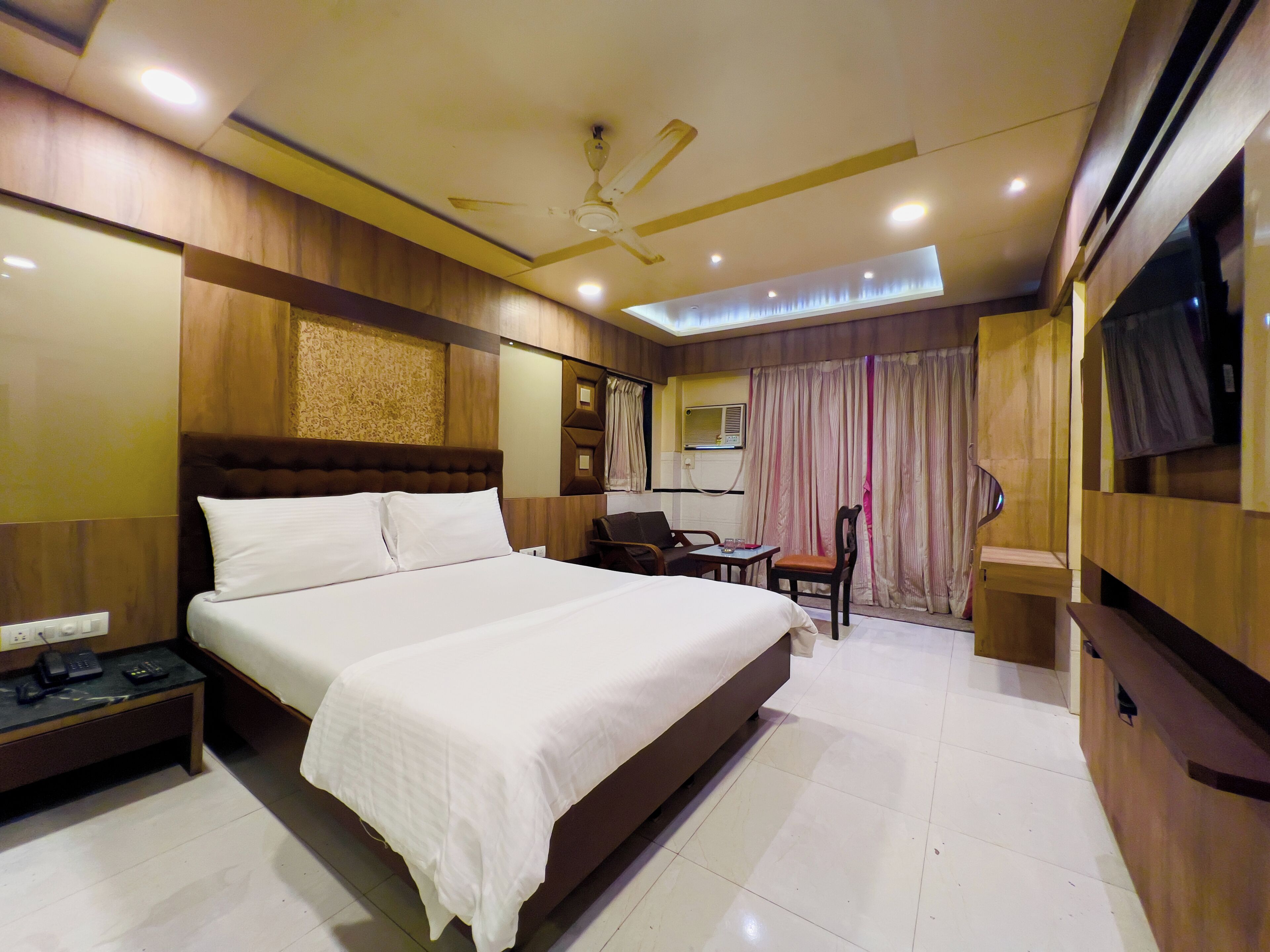 A hotel room with a large bed, a desk, a chair, a TV, and a ceiling fan.