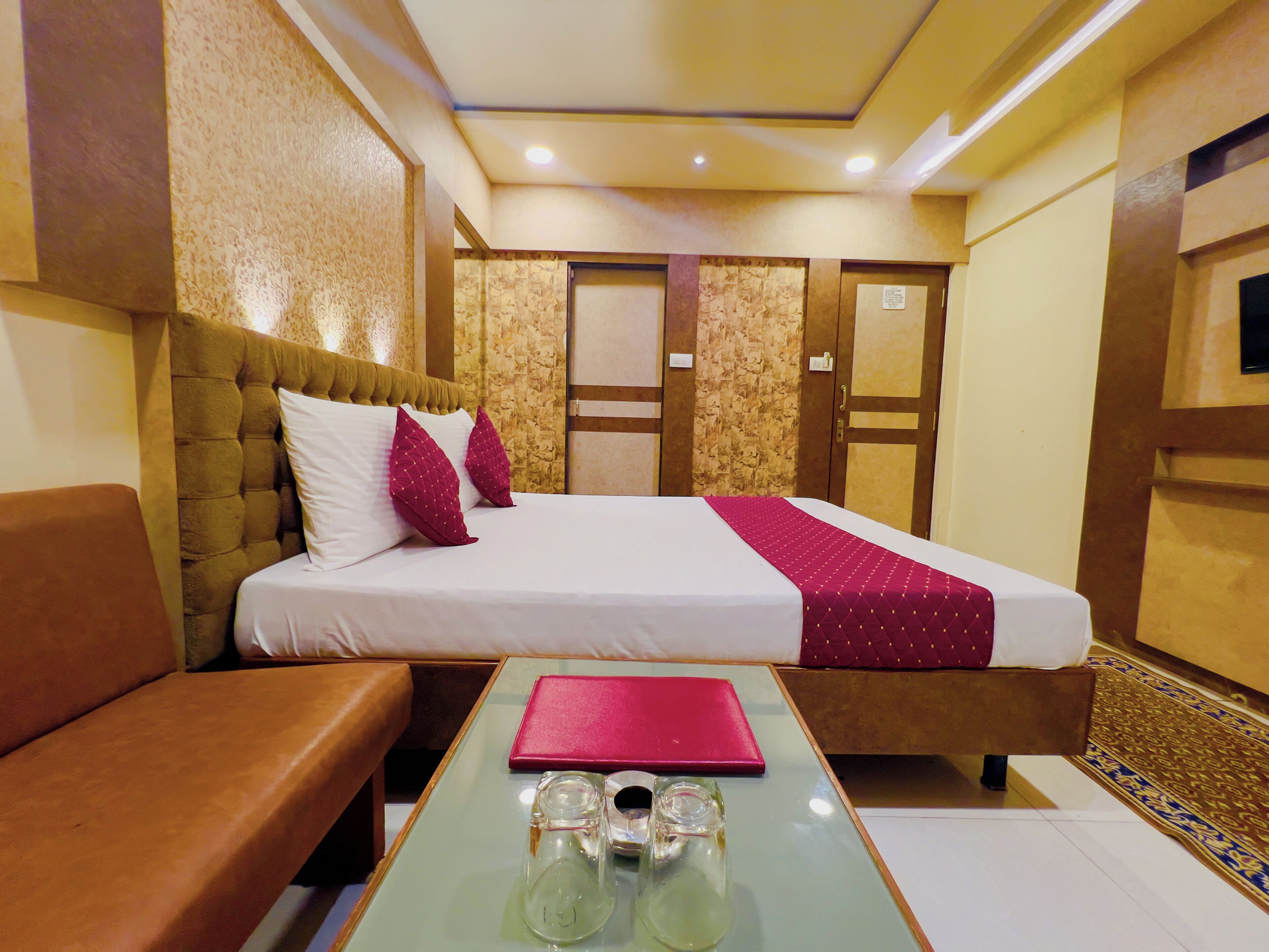 A hotel room with a bed, a sofa, a glass table with a red book and two glasses, and a patterned carpet.
