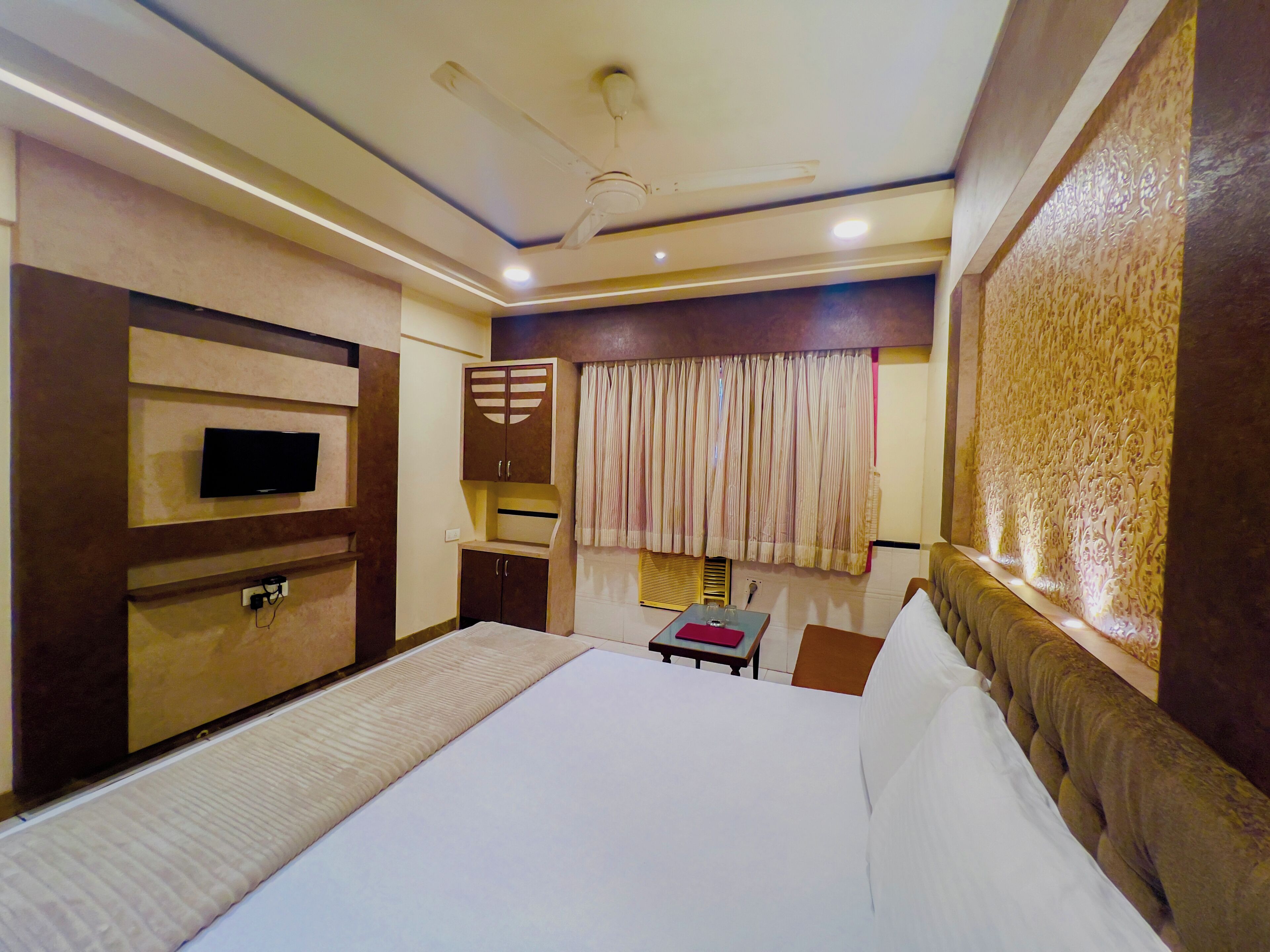 A hotel room with a bed, a TV mounted on the wall, a wardrobe, and a window with curtains.