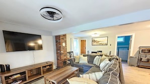 A modern living room with a large flat-screen TV, a wooden coffee table, a grey sofa with striped cushions, and a brick accent wall. - 7 mins to Acadia and walk to town! (Southwest Harbor)