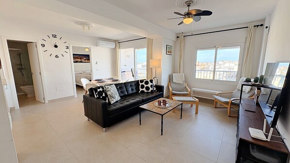 A modern living room with a black leather sofa, a coffee table, and a TV stand with a flat-screen TV. There is a dining area with a table and chairs, and a large window offering a view of the city.