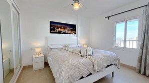 A bedroom with a bed, a nightstand, a lamp, a ceiling fan, a window with curtains, and a cityscape painting on the wall.