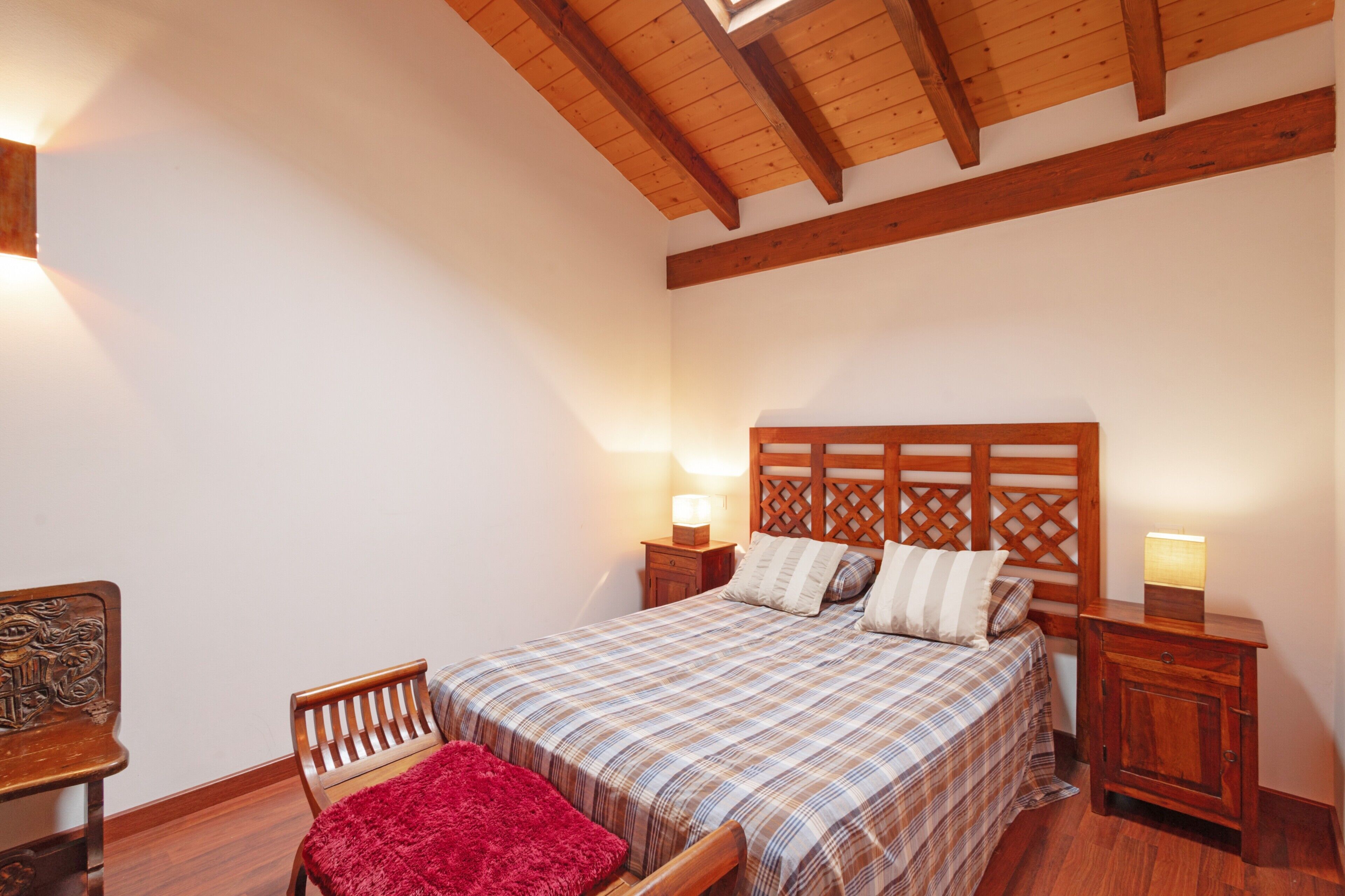 Un dormitorio con cabecera de madera, cama con cubrecama escocés, manta roja, dos mesitas de noche con lámparas y una silla de madera.