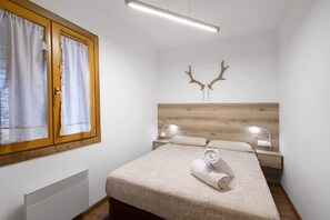 A bedroom with a wooden headboard, a bed with a beige bedspread, a rolled towel on the bed, a nightstand with a lamp, and a window with curtains. - Unique Apartment in Font Romeu (Font-Romeu)