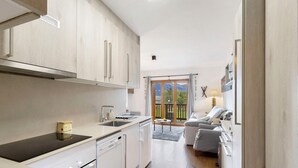 A modern kitchen with a stove, sink, and dishwasher, leading to a living area with a sofa and a view of the outdoors. - Unique Apartment in Font Romeu (Font-Romeu)