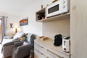 A kitchenette with a microwave, toaster, and coffee maker, a sofa with cushions, a small table, and a wall-mounted shelf with a basket and a picture. - Unique Apartment in Font Romeu (Font-Romeu)