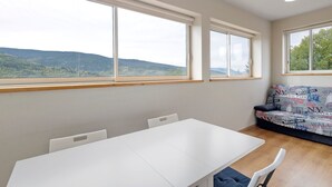 Une pièce avec une table blanche, un canapé orné d’un motif représentant un paysage urbain et de grandes fenêtres donnant sur des collines.