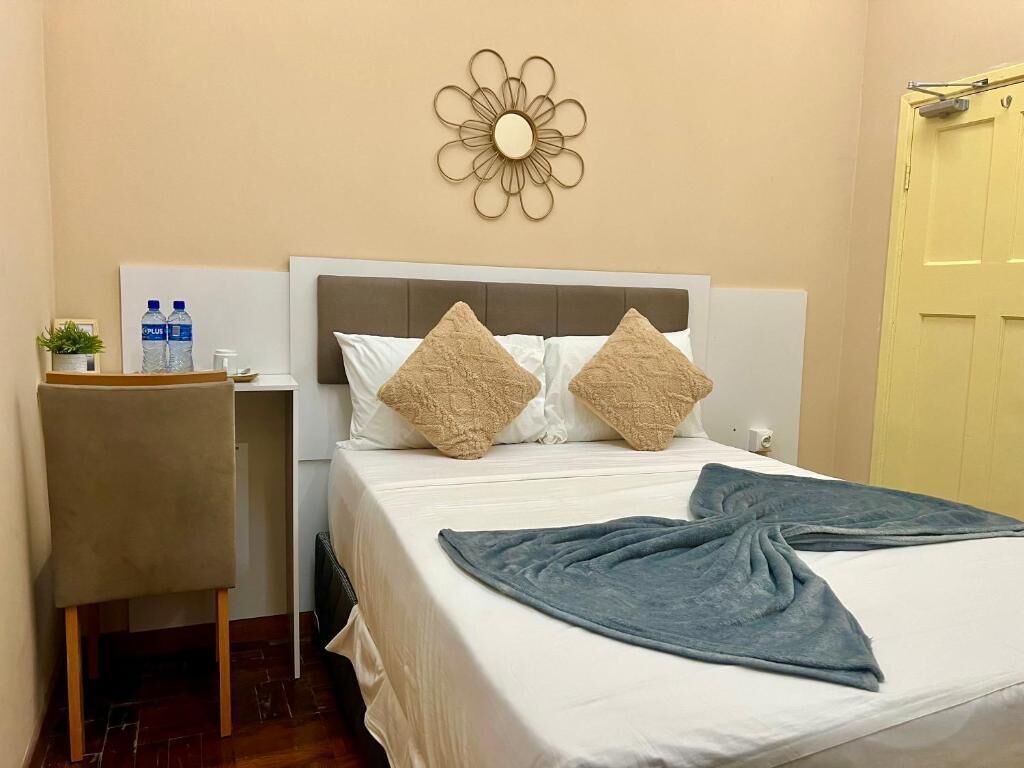 A hotel room with a bed, a headboard, a wall decoration, a desk with a chair, and a door.