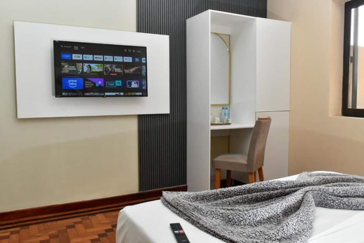 Une chambre d’hôtel équipée d’un téléviseur fixé au mur, qui permet d’accéder à différents services de diffusion en continu, d’un lit avec une couverture, d’une chaise et d’une armoire.
