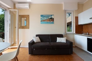Un soggiorno con divano, tavolo da pranzo e sedie. Si nota un poster appeso al muro, una finestra e un angolo cottura con pensili e lavello.
