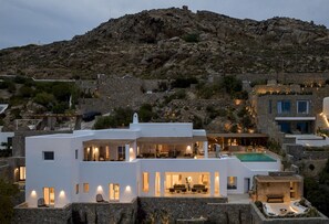 A modern white building with a rooftop pool and outdoor seating area, surrounded by rocky terrain and other structures.