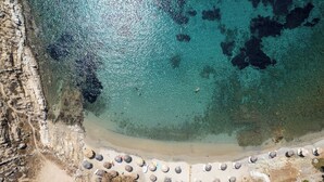 Une vue aérienne d’une plage aux eaux turquoise et limpides, bordée de falaises rocheuses et d’une rangée de parasols.