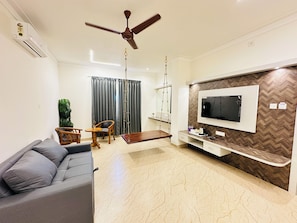 A modern living room with a grey sofa, wooden coffee table, and a TV mounted on a herringbone-patterned wall. - Shambhavi Hotel & Convention Centre (Udupi)