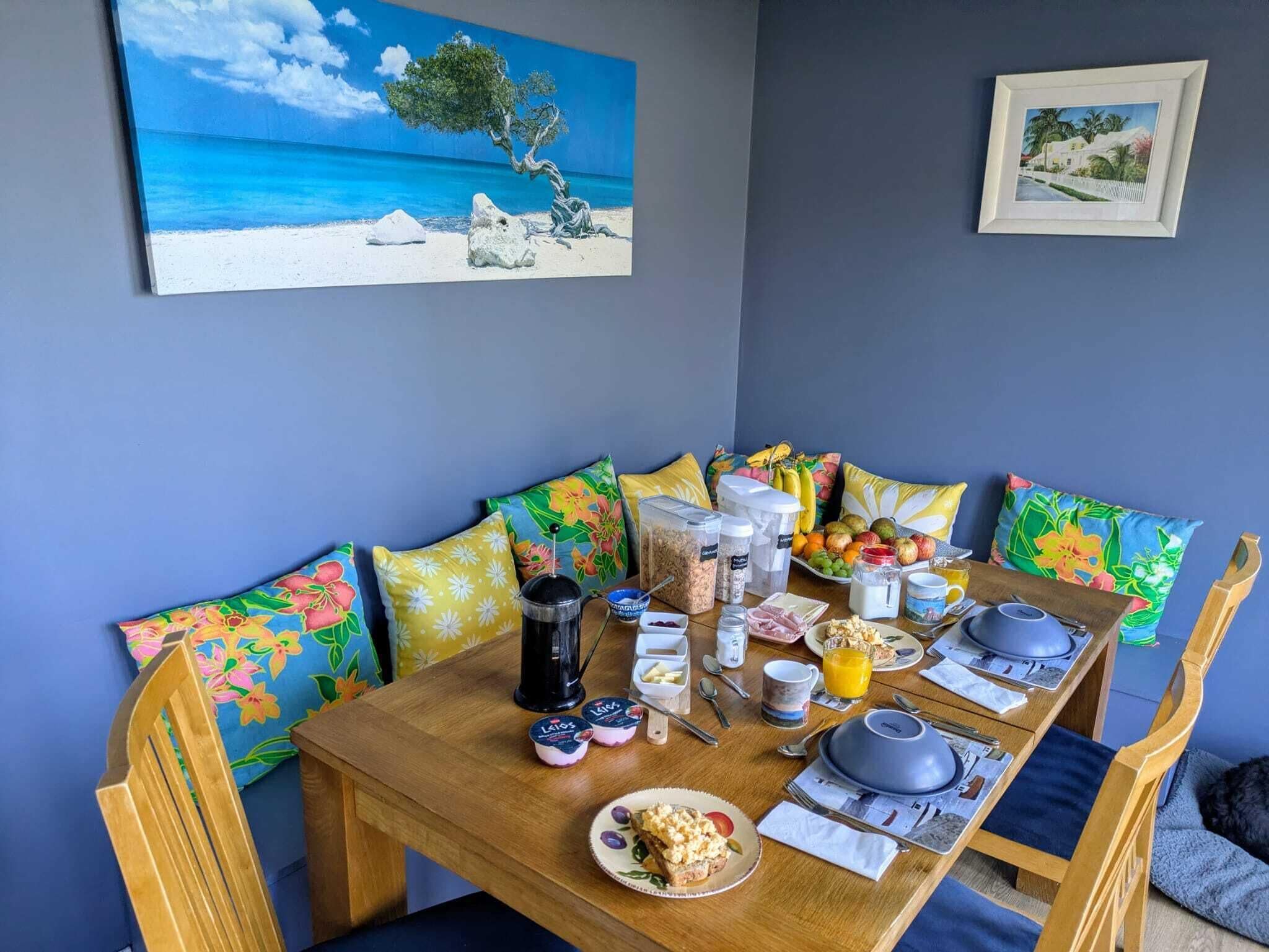 Tavolo apparecchiato per la colazione, circondato da cuscini fioriti colorati, con alle spalle un muro blu e un quadro raffigurante una spiaggia.