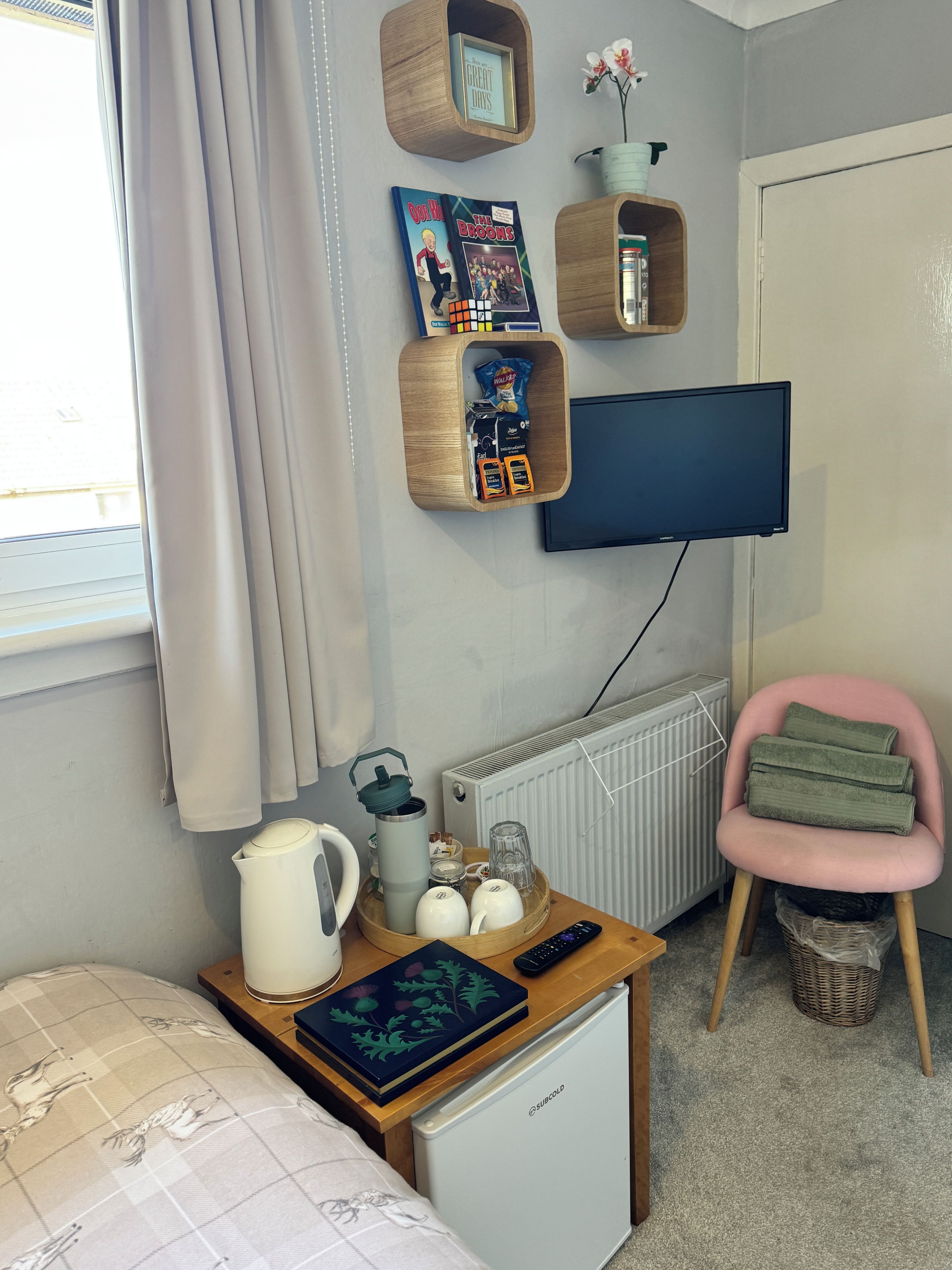Une chambre avec un lit, un petit réfrigérateur, un téléviseur fixé au mur, une étagère en bois avec des livres et un cadre photo, une chaise rose et un radiateur.
