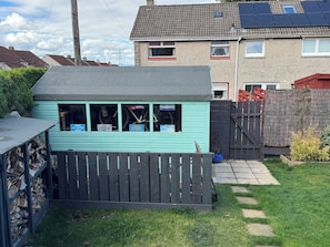 A small, light blue shed with a black roof, a wooden fence, and a stack of firewood to the left. - A perfect location for the airport and the city, comfortable stay home from home (Currie)