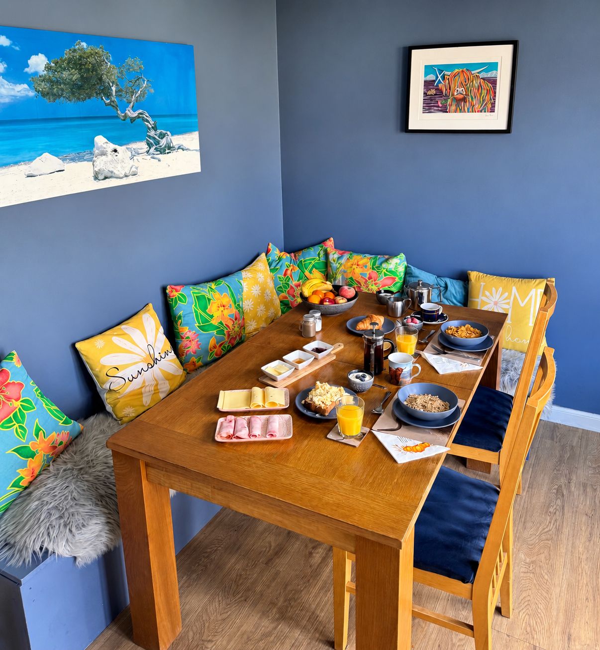 A dining area with a wooden table set for breakfast, colorful pillows, and artwork on the walls.
