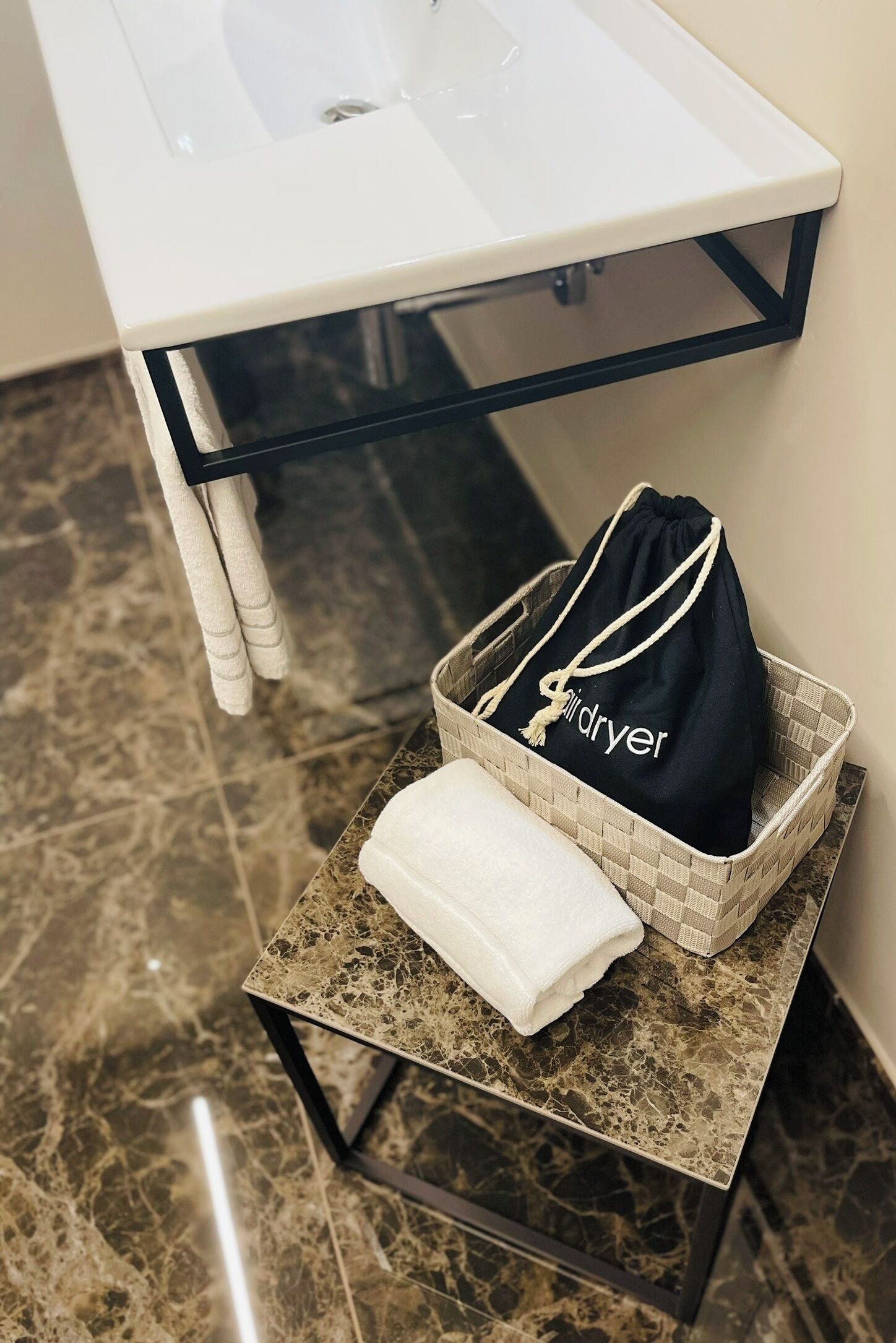 Une salle de bain avec un comptoir en marbre, un lavabo blanc, une étagère en verre et un sac noir étiqueté « sèche-linge » posé sur un panier en osier.