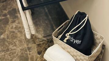 Une salle de bain avec un comptoir en marbre, un lavabo blanc, une étagère en verre et un sac noir étiqueté « sèche-linge » posé sur un panier en osier.