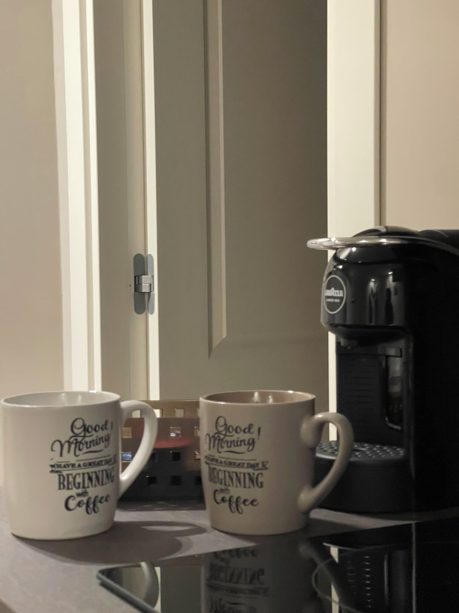 Due tazze bianche con la scritta 'Buongiorno! Che bella giornata per iniziare con il caffè', posate su un tavolo accanto a una macchina da caffè.