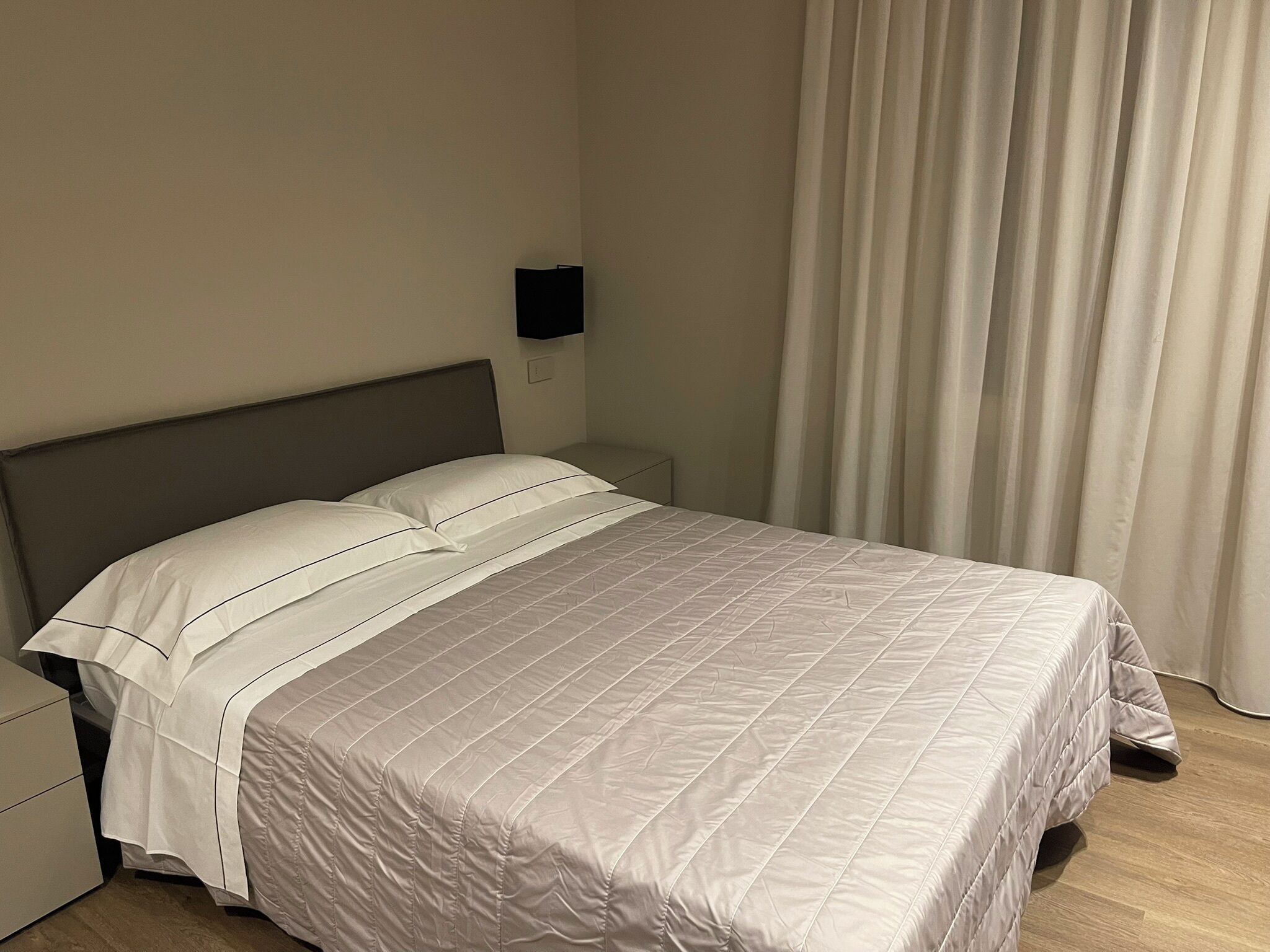 Una cama bien hecha con cubrecama acolchado, una mesita de noche y una lámpara fijada a la pared.