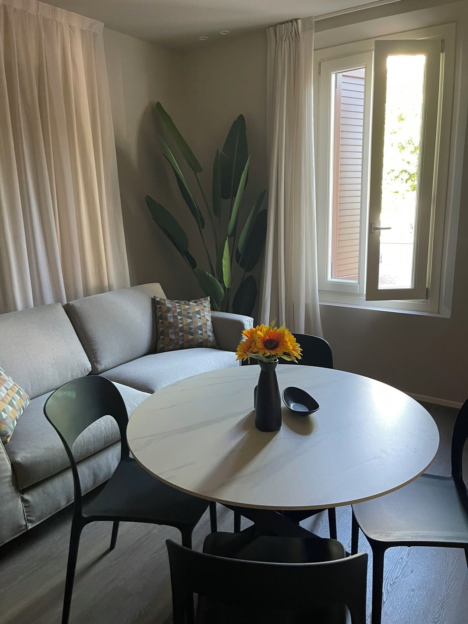 A living room with a white round table, black chairs, a grey sofa, and a large green plant.
