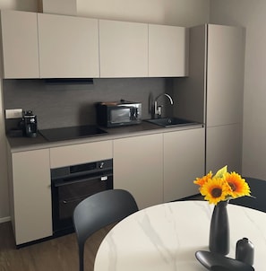 A modern kitchen with a built-in microwave, coffee maker, and sink. There is a dining table with a vase of sunflowers. - La Bella Modena Giardini Appartamenti (Modena)