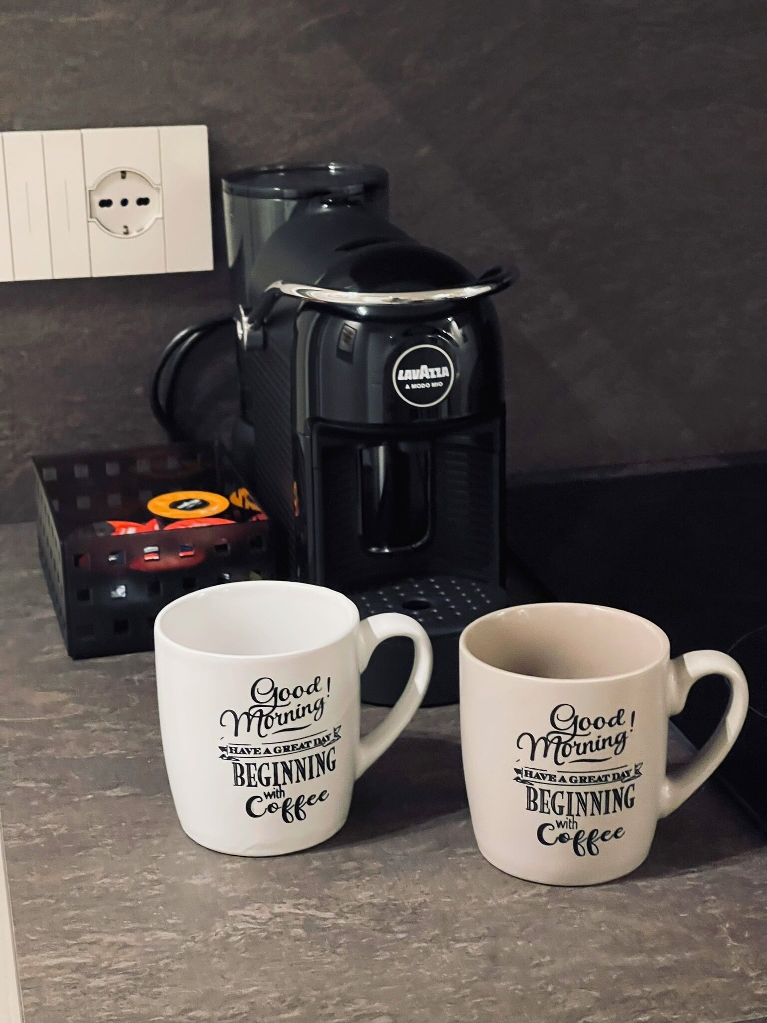 Zwei weiße Kaffeetassen mit dem Aufdruck "Good Morning Have a Great Day Beginning with Coffee" und eine schwarze Kaffeemaschine der Marke "Lavazza" auf einer Küchenarbeitsplatte.