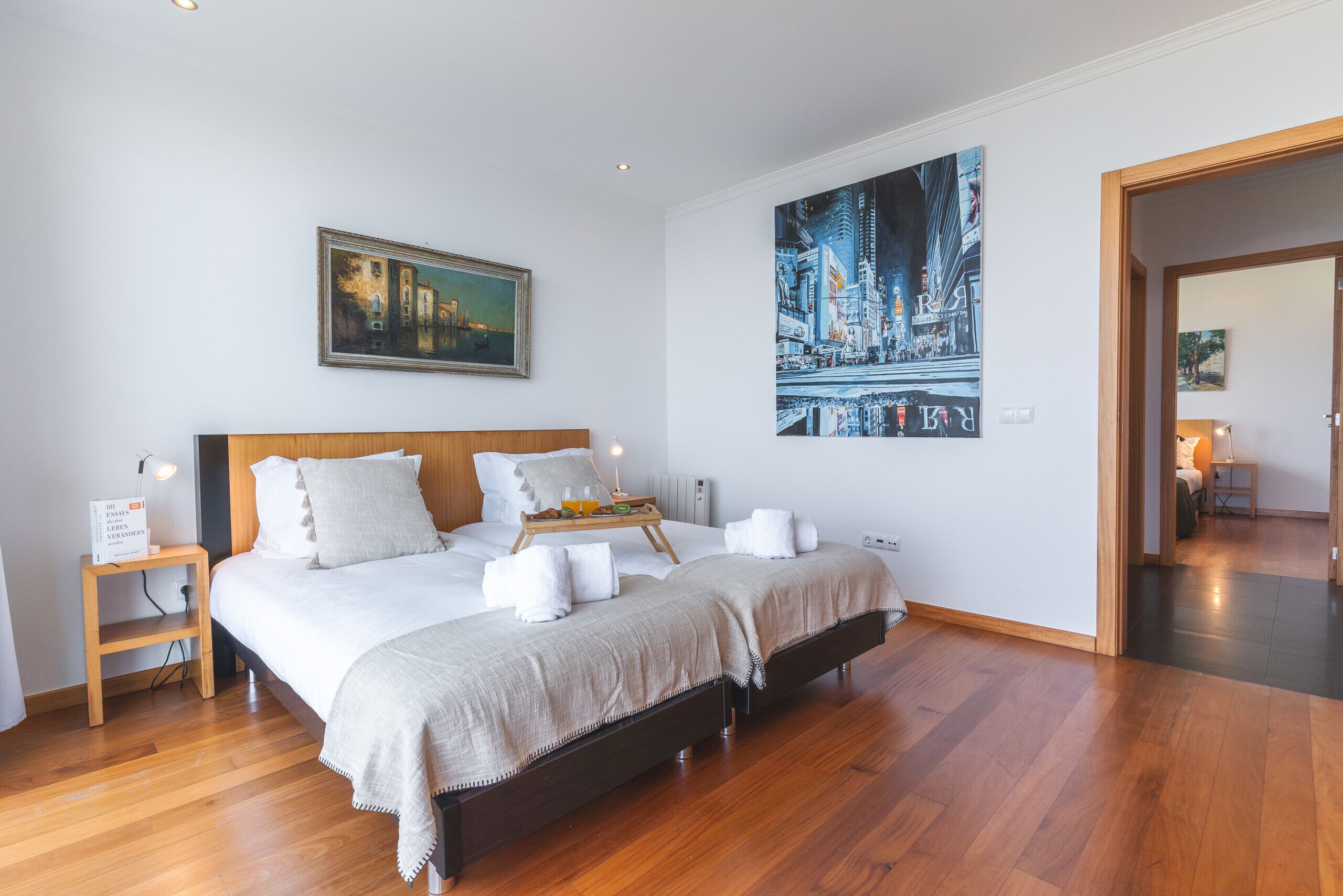 A bedroom with a large bed, a nightstand, a painting, and a cityscape artwork on the wall.