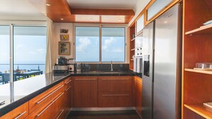 A modern kitchen with wooden cabinets, stainless steel appliances, and a large window overlooking the sea. - Cape View Villa by Atlantic Holiday (Calheta)