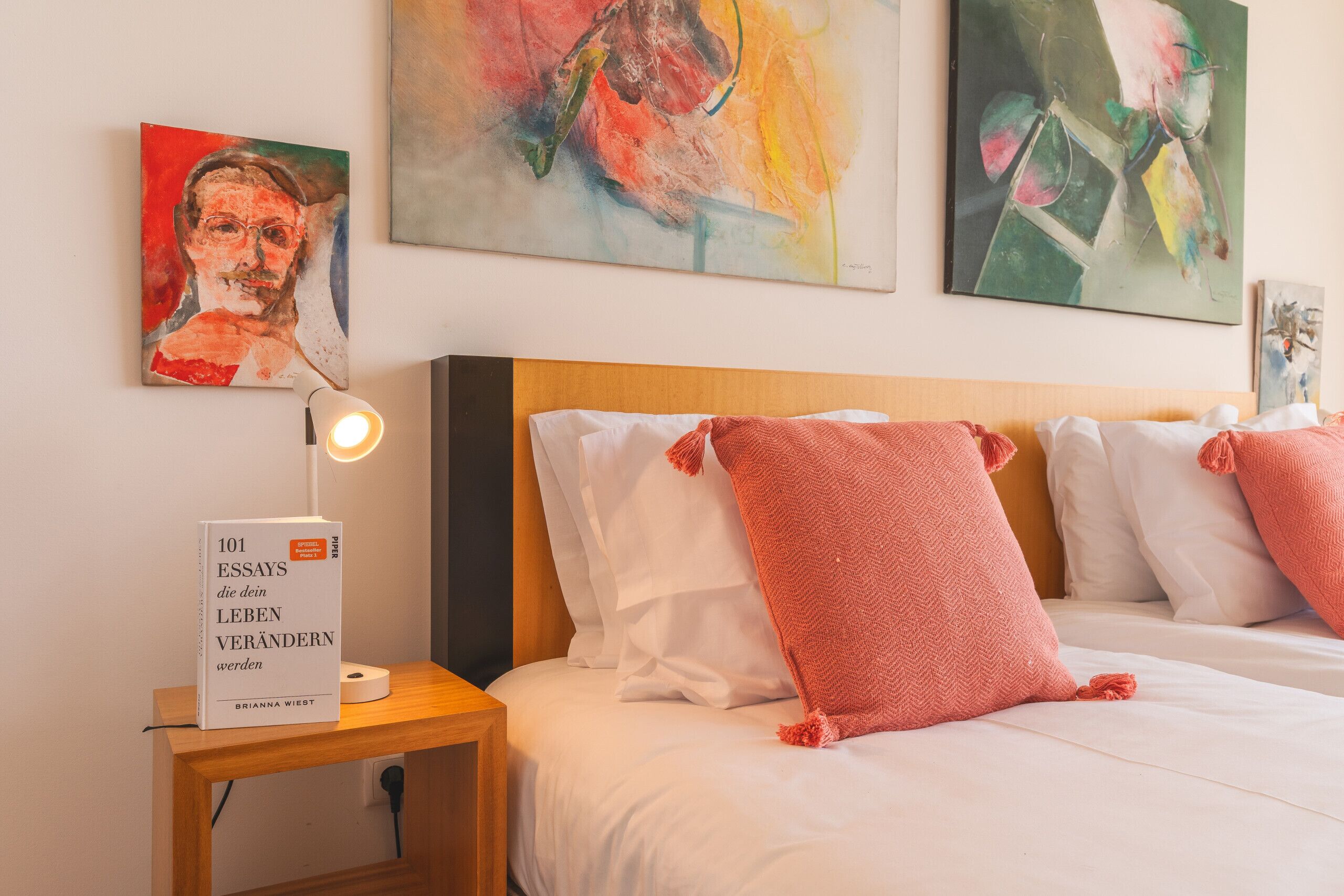 Habitación de hotel con cama, almohadas, mesita de noche con un libro y pinturas abstractas en la pared.
