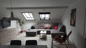 A modern living room with a sectional sofa, a dining table, and a rocking chair. There are skylights on the ceiling and a large window.