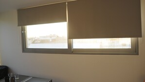 A room with a window covered by a beige roller blind, a small table with a few items, and a view of a building outside. - LOFT WITH PRIVATE TERRACE AND PARTHENON VIEWS (Athina)