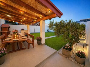 Um pátio externo com mesa de madeira posta para refeições, rodeado por vasos de plantas e uma pequena árvore.