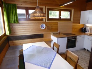 A compact kitchen with wooden walls, a dining table, chairs, a hanging light fixture, and a window with curtains.