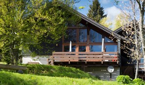 A modern wooden cabin with a balcony, surrounded by lush greenery and trees.