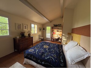 Ein Schlafzimmer mit Bett, Kommode, Bücherregal, Sessel und einem Fenster mit Blick auf Grünflächen.