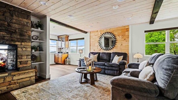 A living room with a stone fireplace, a round mirror, a dark sofa, and a view of the outdoors.