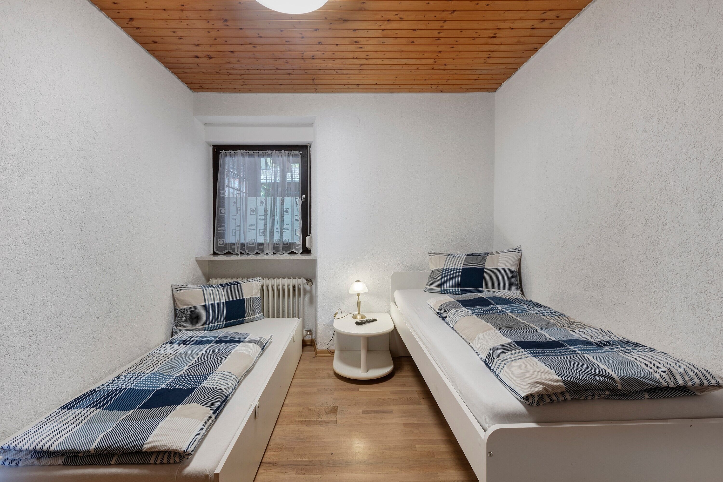 Ein kleiner, gut beleuchteter Raum mit zwei Betten, einem Tisch mit Lampe, einem Fenster mit Vorhängen und einem Heizkörper.