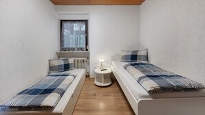 A small, well-lit room with two beds, a small table with a lamp, a window with curtains, and a radiator.