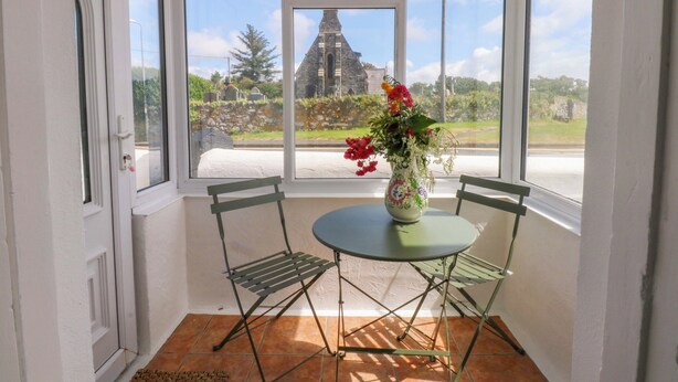 A conservatory with a table and chairs, a vase with flowers, and a view of a church and greenery. - Cae Gwyn (Ty Croes)