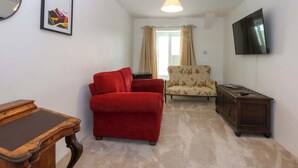 A living room with a red sofa, a floral armchair, a wooden side table, a flat-screen TV, and a ceiling light. - Cae Gwyn (Ty Croes)