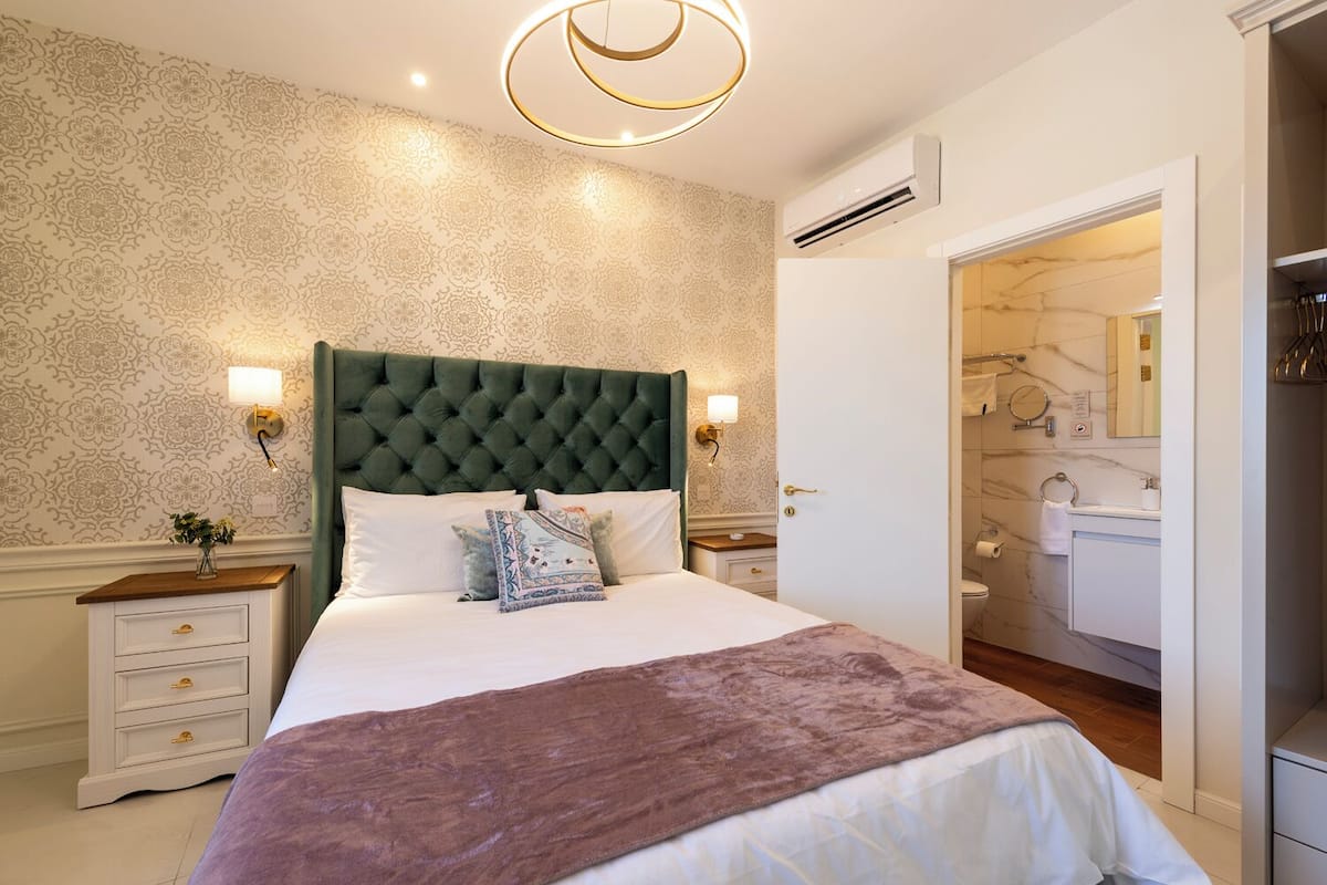 A bedroom with a tufted headboard, a large bed with white linens, two bedside tables with lamps, and a patterned wallpaper.