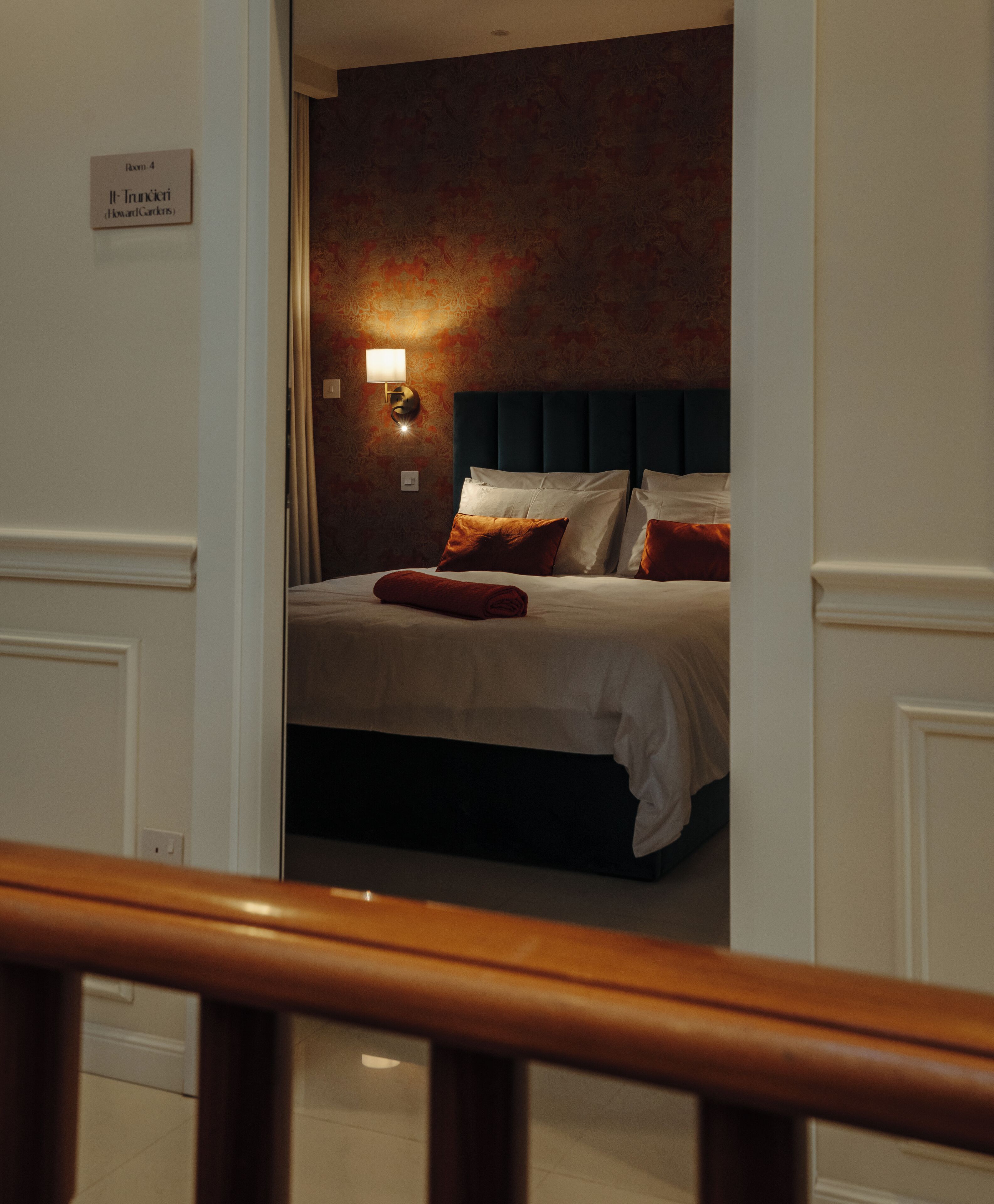 Habitación de hotel con cama, mesita de noche, lámpara de pared y un cartel con el número y nombre de la habitación.