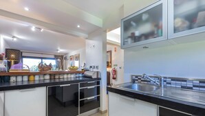 Una cocina moderna con encimera negra, pileta de acero inoxidable y horno empotrado. Hay una ventana con cortinas y un frutero sobre la encimera.