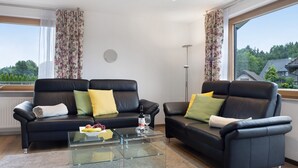 A modern living room with two black leather sofas, a glass coffee table, and a view of a residential area through large windows.
