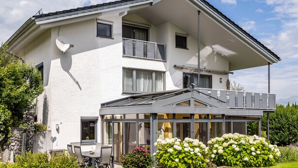 Une maison à deux étages dotée d’un balcon, de grandes fenêtres et d’un jardin où poussent des fleurs blanches.