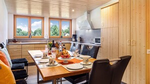 Une cuisine moderne avec des meubles en bois, une table à manger pour quatre personnes et de grandes fenêtres donnant sur un paysage verdoyant.