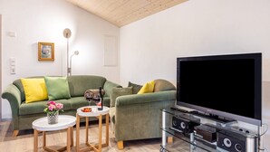 Un salon avec un canapé vert, un téléviseur à écran plat, une table basse en verre et une petite table d’appoint sur laquelle se trouvent un verre à vin et des fleurs.